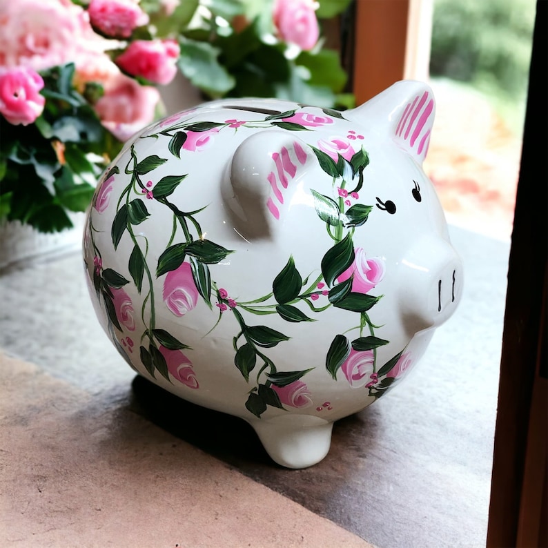 May include: A white ceramic piggy bank featuring a hand-painted floral design of pink roses with green leaves and vines. The pig has pink striped ears, black painted eyes, and a coin slot on its back. The surface is glossy and smooth.
