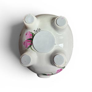 May include: The underside of a white ceramic piggy bank featuring four feet with grey circular pads, a central circular stopper, and hand-painted pink rose motifs with green leaves. The name Nelle is written in black script near one of the feet.