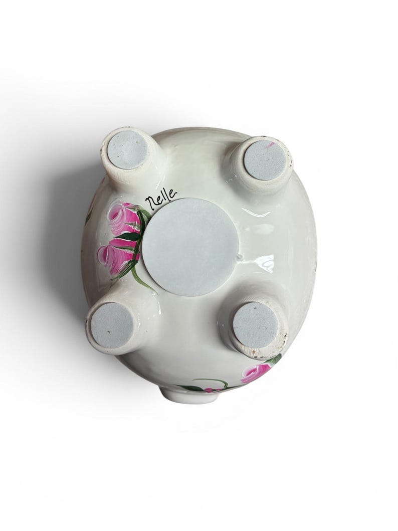May include: The underside of a white ceramic piggy bank featuring four feet with grey circular pads, a central circular stopper, and hand-painted pink rose motifs with green leaves. The name Nelle is written in black script near one of the feet.