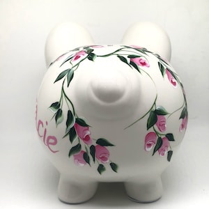 May include: A white ceramic piggy bank featuring a hand-painted floral design of pink rosebuds and green leaves wrapping around the body. The name "Lucie" is painted in pink script on the side. The piggy bank has two ears and a snout.
