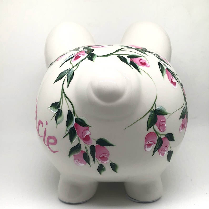 May include: A white ceramic piggy bank featuring a hand-painted floral design of pink rosebuds and green leaves wrapping around the body. The name "Lucie" is painted in pink script on the side. The piggy bank has two ears and a snout.