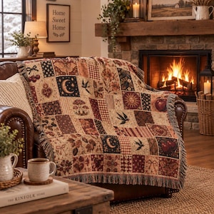 May include: A woven throw blanket featuring a patchwork design of various motifs including cherries, moths, crescent moons, mushrooms, suns, birds, and hearts in shades of red, brown, black, and cream with a fringed edge draped over a leather chair.