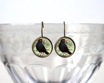 Può includere: Un paio di orecchini pendenti con un merlo nero con becco arancione su sfondo verde. Gli orecchini sono in metallo con finitura bronzo.