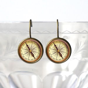Puede incluir: Un par de pendientes de brújula de color dorado con una cara blanca y una aguja roja. La cara de la brújula tiene marcas negras y una estrella negra en el centro.