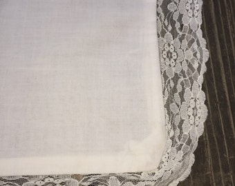 Pode incluir: Quadrado de tecido branco com uma borda de renda branca ao redor das bordas. O tecido é liso e a renda é delicada.