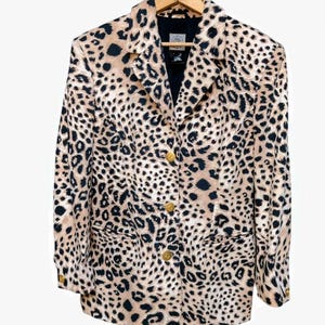May include: A tan and black leopard print blazer with gold buttons. The blazer is hanging on a wooden hanger. The jacket has a classic collar, two front pockets, and a black lining.