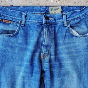 Puede incluir: Un par de pantalones vaqueros azules de mezclilla con una etiqueta Wrangler de cuero marr&oacute;n en el bolsillo trasero. Los vaqueros tienen un estilo cl&aacute;sico de cinco bolsillos y cierre de bot&oacute;n.