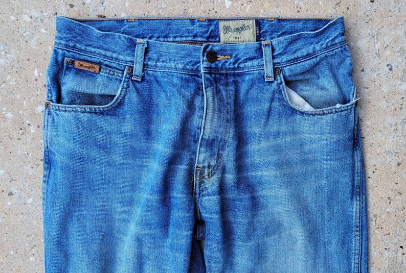 Puede incluir: Un par de pantalones vaqueros azules de mezclilla con una etiqueta Wrangler de cuero marr&oacute;n en el bolsillo trasero. Los vaqueros tienen un estilo cl&aacute;sico de cinco bolsillos y cierre de bot&oacute;n.
