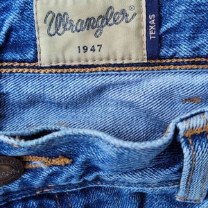 Puede incluir: Primer plano de un bolsillo de mezclilla azul con un parche de cuero marr&oacute;n que dice "Wrangler 1947" y un parche negro m&aacute;s peque&ntilde;o que dice "Texas".