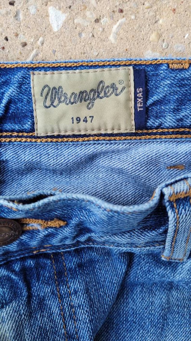 Puede incluir: Primer plano de un bolsillo de mezclilla azul con un parche de cuero marr&oacute;n que dice "Wrangler 1947" y un parche negro m&aacute;s peque&ntilde;o que dice "Texas".