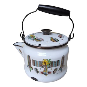 May include: A white enamel tea kettle featuring a black handle and a lid with a dark knob. The body is decorated with colorful graphic illustrations of corn stalks and abstract shapes. The rim and spout edges are trimmed in a dark brown metal finish.