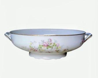 May include: A white porcelain serving bowl featuring a delicate floral pattern of pink roses and green leaves around the exterior. The bowl has two small handles on opposite sides, a scalloped base, and a thin gold rim along the top edge.