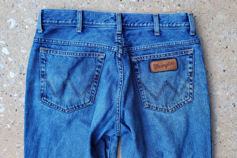 Puede incluir: Un par de pantalones vaqueros azules con un parche de cuero marr&oacute;n que dice "Wrangler" en el bolsillo trasero.