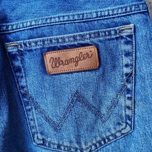 Puede incluir: Primer plano de un bolsillo trasero de mezclilla azul con un parche de cuero marr&oacute;n que dice "Wrangler" en letras blancas.