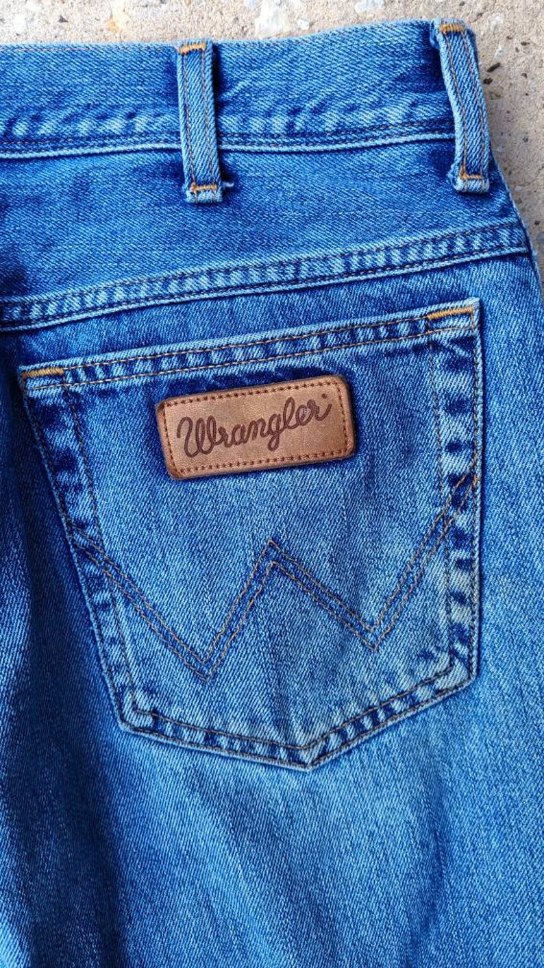 Puede incluir: Primer plano de un bolsillo trasero de mezclilla azul con un parche de cuero marr&oacute;n que dice "Wrangler" en letras blancas.