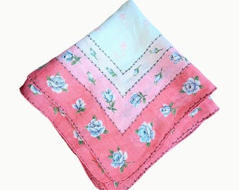 Pode incluir: Um lenço quadrado dobrado com um padrão floral. O lenço apresenta um gradiente de cores, passando do rosa claro a um tom rosa mais profundo. Rosas azuis estão espalhadas pelo tecido, com uma borda delicada.