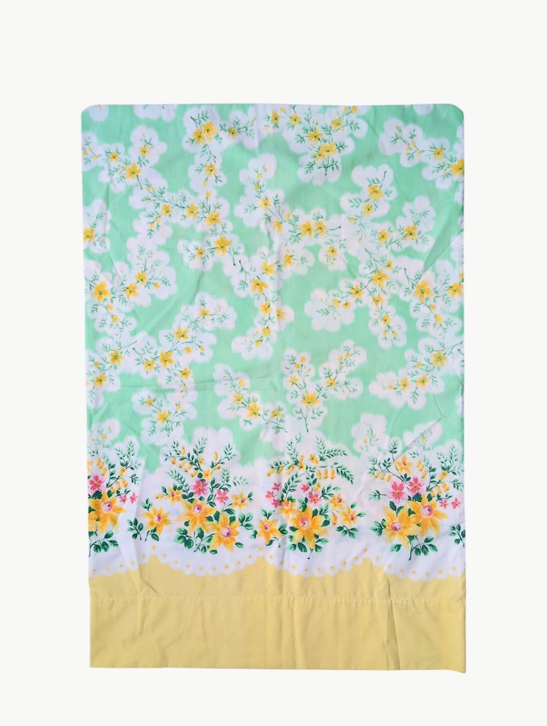 Pode incluir: Um len&ccedil;ol retangular com estampa floral. O fundo &eacute; verde claro, com flores brancas, amarelas e rosas. A borda inferior &eacute; amarela com uma borda ondulada branca.