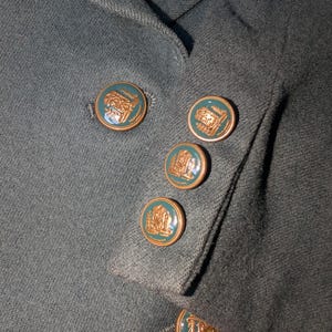 May include: Close-up of a dark teal blazer with decorative gold-colored buttons. The buttons have a teal center with a gold-colored emblem. The blazer's fabric has a textured appearance.