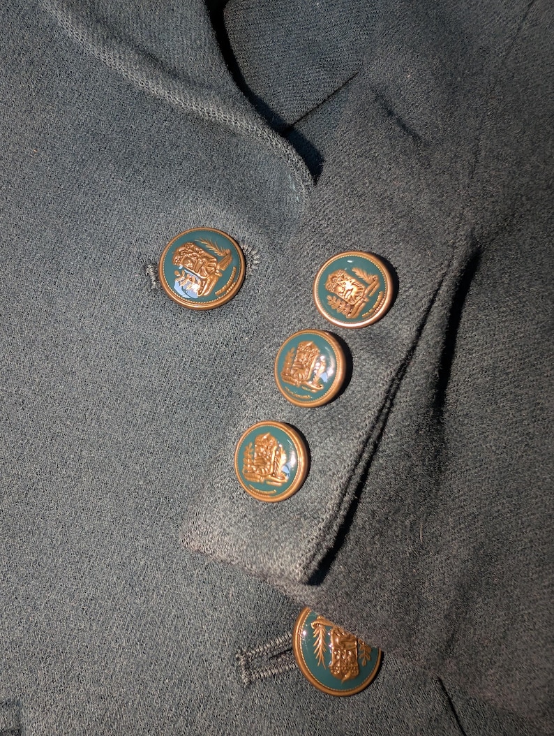 May include: Close-up of a dark teal blazer with decorative gold-colored buttons. The buttons have a teal center with a gold-colored emblem. The blazer's fabric has a textured appearance.