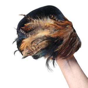 May include: A black hat adorned with a dense cluster of layered feathers in shades of brown, tan, and black. The feathers are arranged in a textured, overlapping pattern across the side of the dark, smooth-surfaced headwear piece.