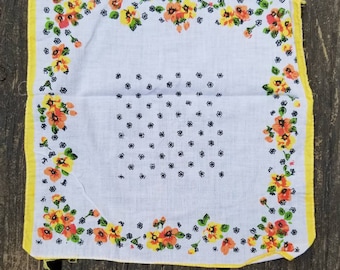 Pode incluir: Um lenço de algodão branco com uma borda amarela e um padrão floral. As flores são laranja, amarelo e vermelho com folhas verdes. O centro do lenço tem um padrão de pequenos pontos pretos.
