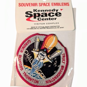 Könnte beinhalten: Ein Souvenir-Weltraum-Emblem vom Kennedy Space Center. Der Aufnäher zeigt ein Raumschiffdesign mit den Namen Gardner, Bluford, Thornton und Brandenstein. Der Aufnäher befindet sich in einer transparenten Plastikhülle mit einem weißen Header mit der Aufschrift "SOUVENIR SPACE EMBLEMS".