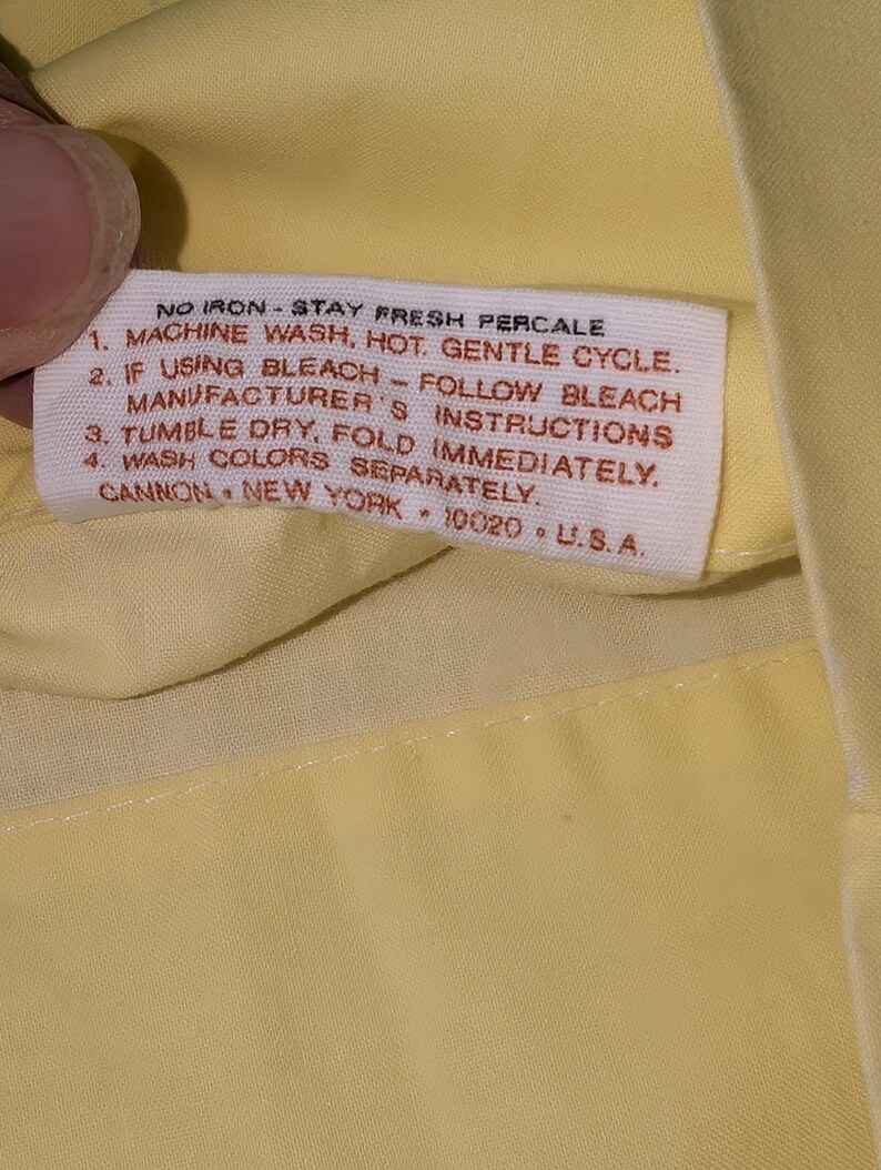 Pode incluir: Close-up de um tecido amarelo com uma etiqueta branca. A etiqueta tem o texto "NO IRON - STAY FRESH PERCALE" e instru&ccedil;&otilde;es de lavagem. A etiqueta tamb&eacute;m inclui a marca "CANNON, NEW YORK, 10020, U.S.A."