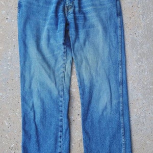 Puede incluir: Un par de pantalones vaqueros azules con un parche de cuero marr&oacute;n que dice "Wrangler" en la parte posterior de la cintura.