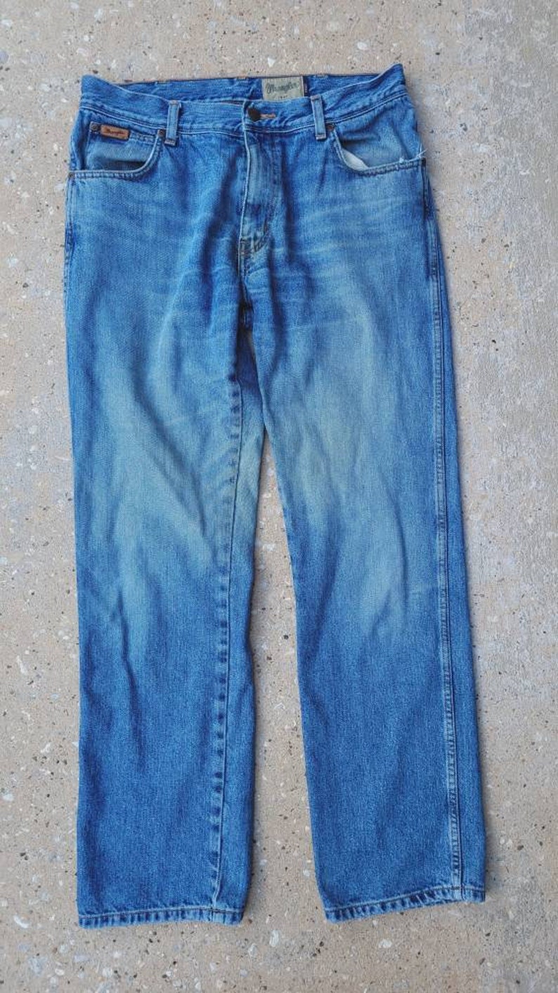 Puede incluir: Un par de pantalones vaqueros azules con un parche de cuero marr&oacute;n que dice "Wrangler" en la parte posterior de la cintura.