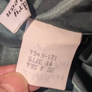 May include: Close-up of a garment tag with the text "dry clean" on a white label. The tag also includes the size "16" and other numbers. The fabric is a dark green color.