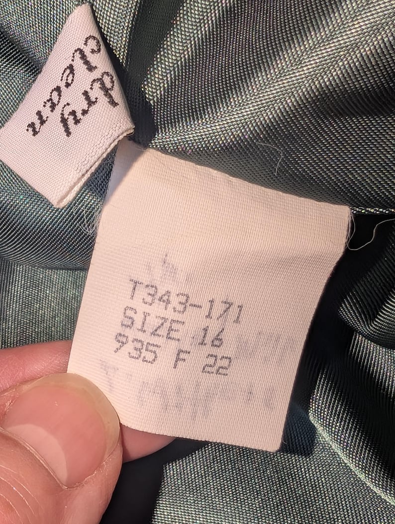 May include: Close-up of a garment tag with the text "dry clean" on a white label. The tag also includes the size "16" and other numbers. The fabric is a dark green color.