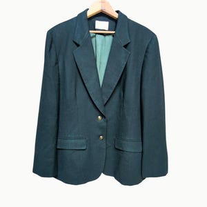May include: A dark green blazer with a notched lapel, two front pockets, and gold-colored buttons. The blazer is hanging on a wooden hanger against a white background. The interior lining is a lighter green color.