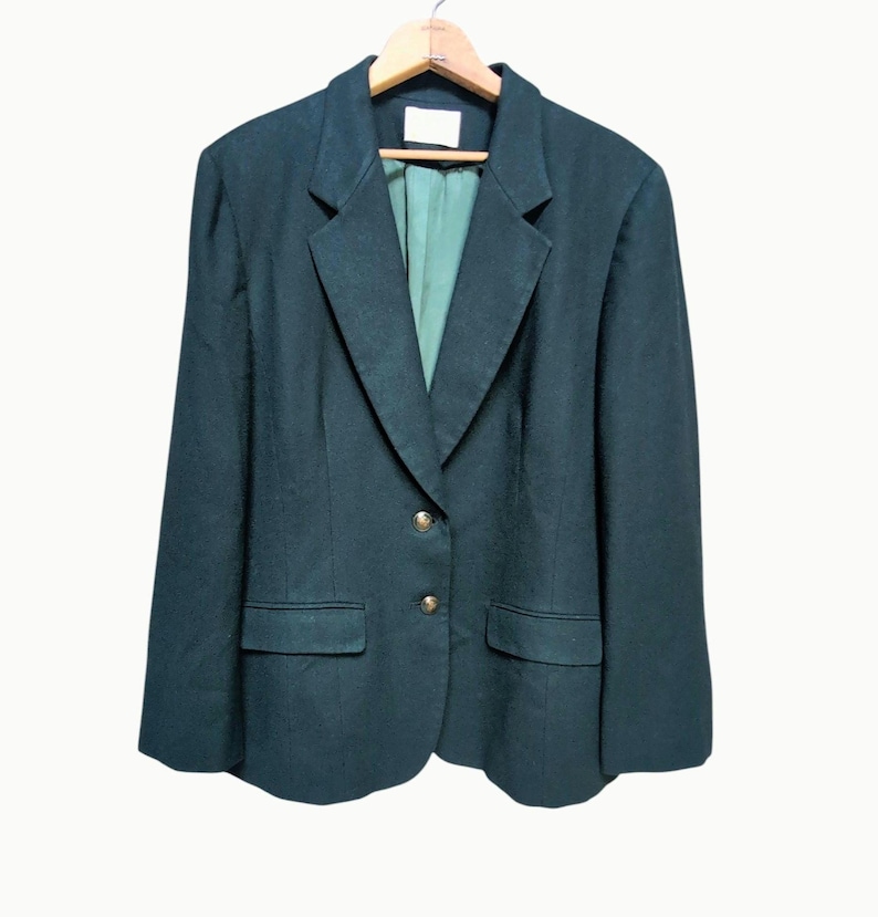 May include: A dark green blazer with a notched lapel, two front pockets, and gold-colored buttons. The blazer is hanging on a wooden hanger against a white background. The interior lining is a lighter green color.