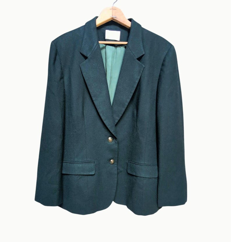 May include: Dark green blazer with a notched lapel, two front pockets, and gold-colored buttons. The blazer is hanging on a wooden hanger. The interior lining is a lighter shade of green.