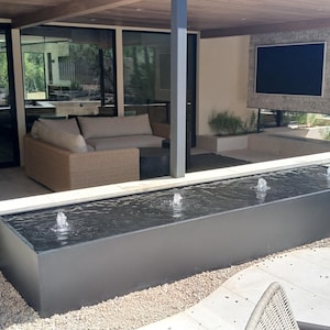 May include: A modern outdoor patio with a rectangular water fountain. The fountain has three water jets. A beige sofa and a large flat-screen TV are visible. The patio is surrounded by glass doors and windows.