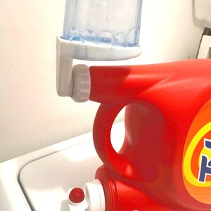 May include: A red laundry detergent bottle with a white and blue measuring cup attached. The bottle has a white spout and a red cap. The measuring cup is clear with a blue top.