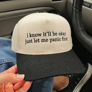 May include: A beige and black baseball cap with the text "i know it'll be okay just let me panic first" in black lettering. The cap is being held in a person's hand.