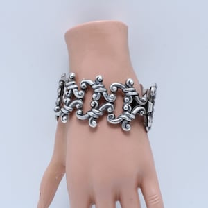 Op de afbeelding: Een zilveren armband met een gedetailleerd, ingewikkeld ontwerp. De armband is gemaakt van met elkaar verbonden, gedraaide en gebogen metalen stukken.