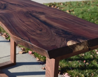 May include: A dark brown wooden table with a live edge top and simple, modern legs. The table is made of walnut wood and has a natural, rustic finish.