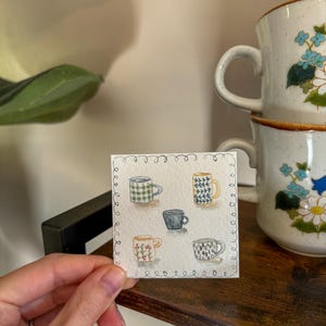 May include: A watercolor painting of six small coffee mugs with various patterns, including plaid, floral, and geometric designs. Two larger, stacked mugs with blue floral patterns are in the background. The painting is held by a hand on a wooden surface.