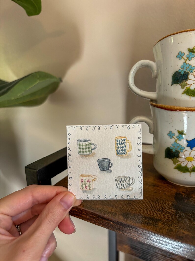 May include: A watercolor painting of six small coffee mugs with various patterns, including plaid, floral, and geometric designs. Two larger, stacked mugs with blue floral patterns are in the background. The painting is held by a hand on a wooden surface.
