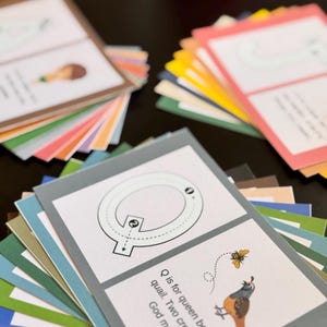 Puede incluir: Una colección de tarjetas didácticas coloridas, cada una con una letra del alfabeto. Las tarjetas están dispuestas en pilas, con la tarjeta de la letra "Q" en el centro, mostrando una línea punteada para trazar y una ilustración de una codorniz y una abeja. Las tarjetas tienen una variedad de colores.
