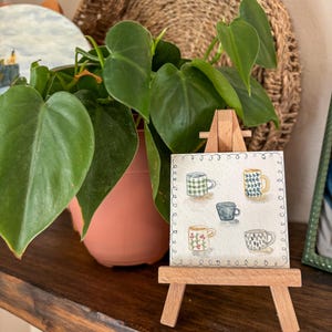 May include: A small wooden easel displays a watercolor painting of five patterned mugs. The mugs are in various colors and designs, including green plaid, blue and yellow geometric, and floral patterns. A potted plant with green leaves is in the background.