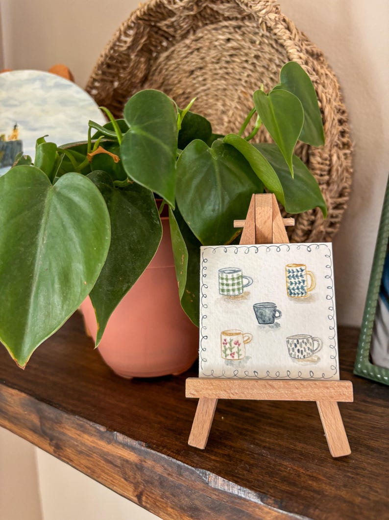 May include: A small wooden easel displays a watercolor painting of five patterned mugs. The mugs are in various colors and designs, including green plaid, blue and yellow geometric, and floral patterns. A potted plant with green leaves is in the background.