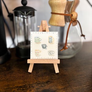 May include: A small wooden easel displays a watercolor painting of six different coffee mugs. The mugs are in various colors and patterns, including green gingham, blue and white geometric designs, and floral motifs. A French press and a pour-over coffee maker are in the background.