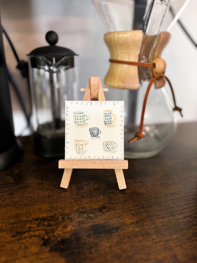 May include: A small wooden easel displays a watercolor painting of six different coffee mugs. The mugs are in various colors and patterns, including green gingham, blue and white geometric designs, and floral motifs. A French press and a pour-over coffee maker are in the background.