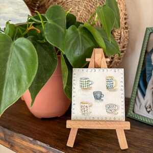 May include: A small wooden easel displays a watercolor painting of five different coffee mugs. The mugs are decorated with various patterns, including checks, floral designs, and geometric shapes. A green plant in a terracotta pot sits to the left of the easel.