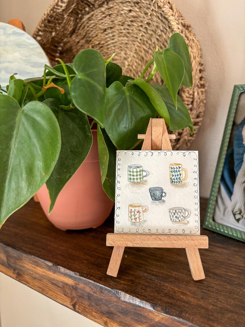 May include: A small wooden easel displays a watercolor painting of five different coffee mugs. The mugs are decorated with various patterns, including checks, floral designs, and geometric shapes. A green plant in a terracotta pot sits to the left of the easel.