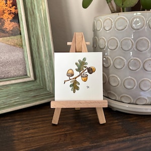 Puede incluir: Una pequeña pintura de acuarela de bellotas y hojas de roble sobre un caballete de madera en miniatura. La obra de arte presenta tres bellotas en tonos marrones y dorados, con hojas verdes. El caballete se exhibe sobre una superficie de madera oscura.