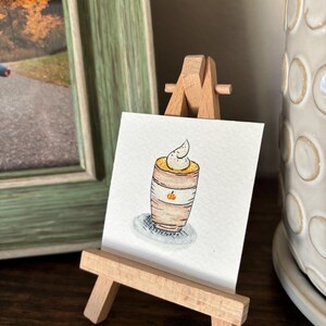 Puede incluir: Una acuarela de un café con leche de calabaza en un pequeño caballete de madera. El café con leche está en una taza marrón con crema batida blanca y una pequeña ilustración de calabaza. El caballete está sobre una superficie de madera oscura.